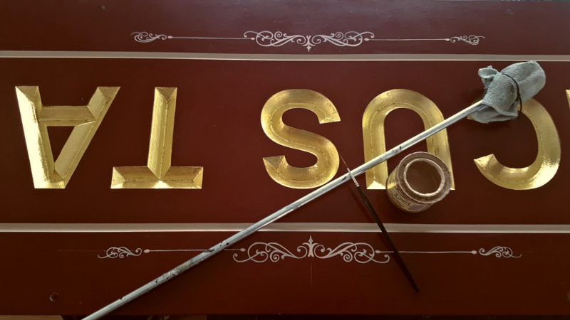 Ivory scroll embellishments being painted onto the gold leaf wooden sign using a specialist signwriting brush.