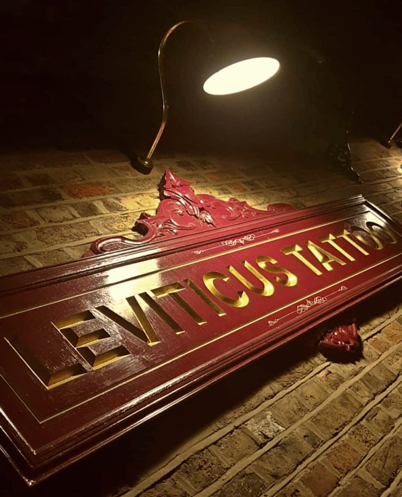 Gold leaf wooden sign for Leviticus Tattoo illuminated by a swan-neck lamp at dusk in Loughton.
