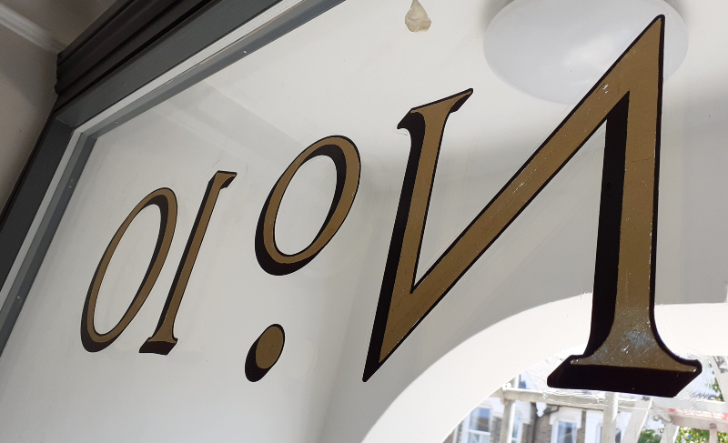 Repainting the outline and cast shadow behind reverse glass gold leaf property numbering to achieve a consistent appearance from both sides of the glazing.