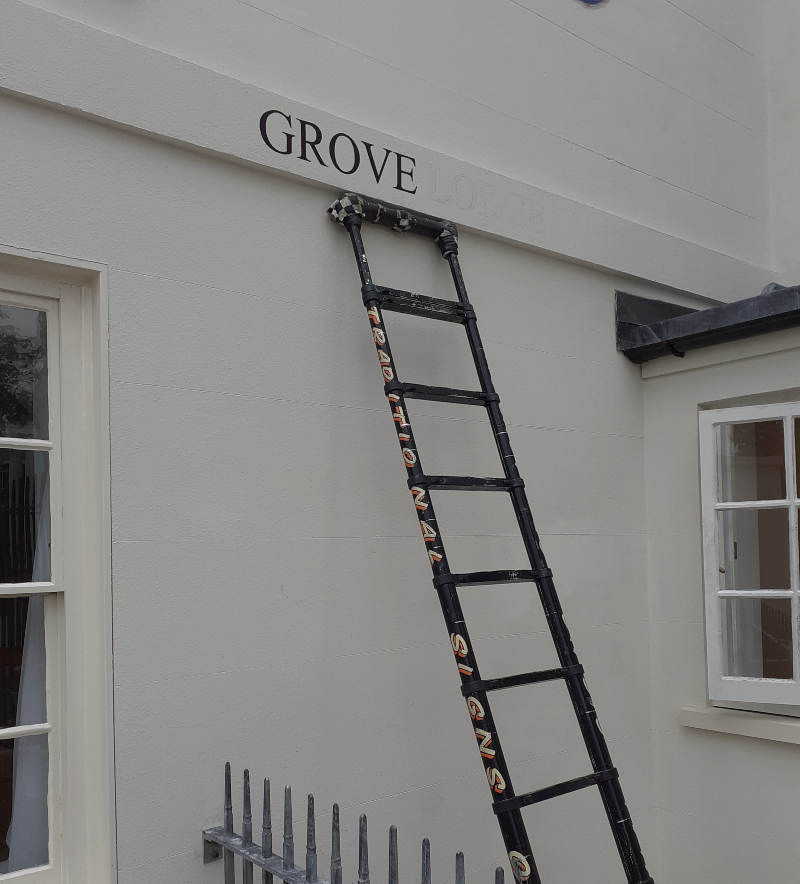 Traditional signwriter painting the Grove Lodge property name directly onto a stippled masonry lintel using signwriting enamel.